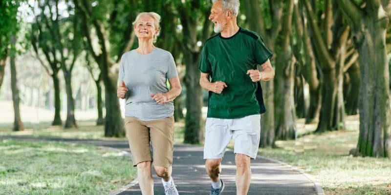 Mature white woman and man jog along path through cluster of trees as they chat.