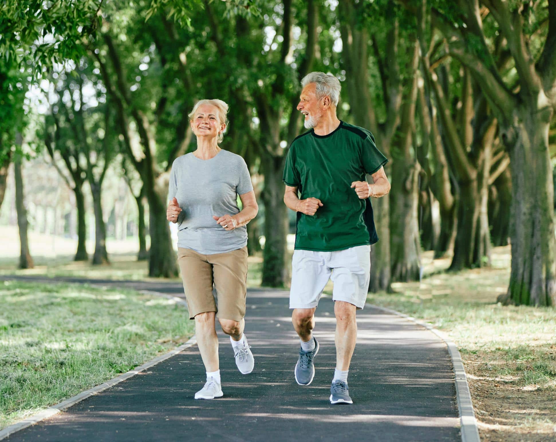 Mature white woman and man jog along path through cluster of trees as they chat.