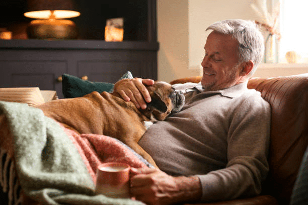Older man lying on the couch smiling at his dog lying on his chest while holding a cup of coffee.